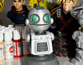 May include: A silver robot figurine with green eye-like lenses and a red antenna. The robot is on a gray circular base. Other anime character busts and paint bottles are in the background.