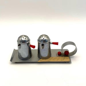 May include: A vintage chrome salt and pepper shaker set with red handles, resting on a rectangular metal tray. The set includes two shakers, a small ring, and two red spheres. A small piece of paper is also on the tray.