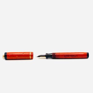 Może przedstawiać: Zabytkowe pióro wieczne z czerwonym, marmurkowym korpusem i nasadką, rozdzielone. Pióro posiada czarną stalówkę i złoty pasek na nasadce. Na korpusie pióra znajduje się napis "WRITE RIGHT".