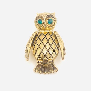 May include: A gold-colored owl brooch with teal-colored rhinestone eyes. The owl's body features a diamond pattern, and its wings are slightly spread. The brooch is a vintage-style accessory.
