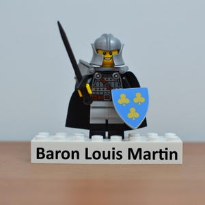 May include: A Lego knight figure with a silver helmet, armor, and a black cape. The knight holds a black sword and a blue shield with yellow clover symbols. The base reads "Baron Louis Martin".