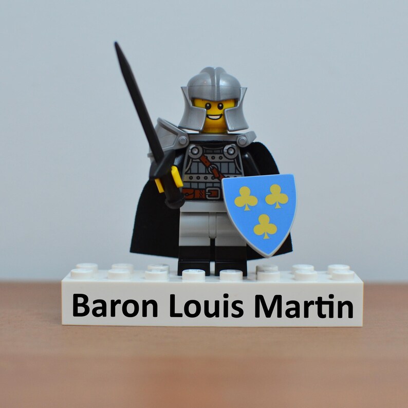 May include: A Lego knight figure with a silver helmet, armor, and a black cape. The knight holds a black sword and a blue shield with yellow clover symbols. The base reads "Baron Louis Martin".