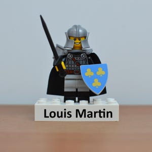 May include: A Lego knight figurine with a silver helmet, armor, and a black cape. The knight holds a black sword and a blue shield with yellow clover symbols. The base of the figurine reads "Louis Martin".