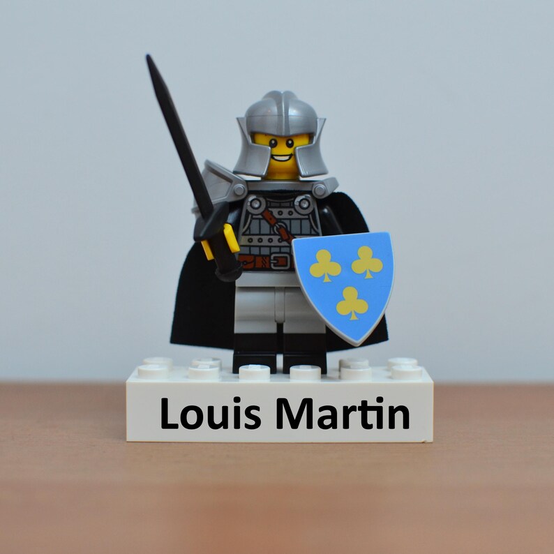 May include: A Lego knight figurine with a silver helmet, armor, and a black cape. The knight holds a black sword and a blue shield with yellow clover symbols. The base of the figurine reads "Louis Martin".