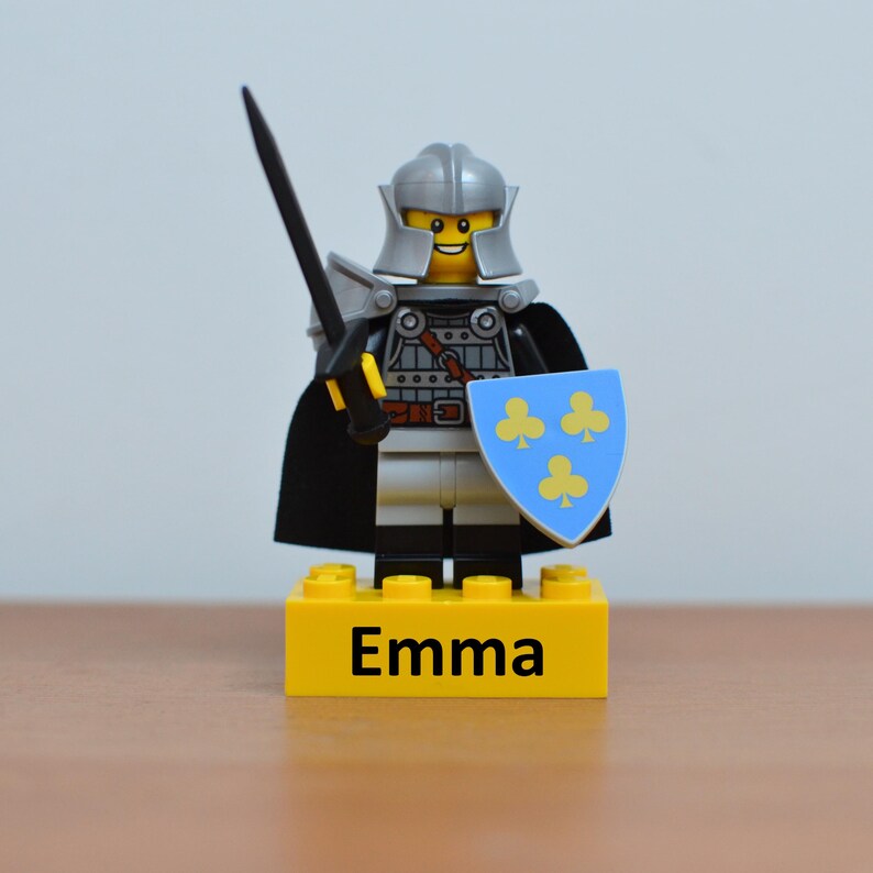 May include: A Lego knight figurine with a silver helmet, black cape, and shield with yellow clover designs. The knight holds a black sword and stands on a yellow base with the name "Emma" printed in black.