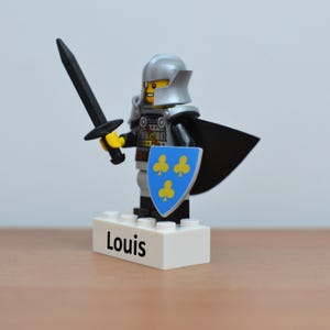 May include: A Lego knight figurine with a silver helmet, black cape, and shield with yellow clover symbols. The knight holds a black sword and stands on a white base with the name "Louis" printed on it.
