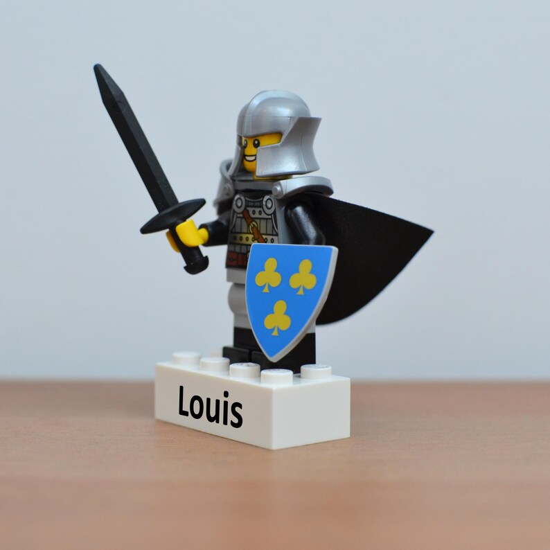 May include: A Lego knight figurine with a silver helmet, black cape, and shield with yellow clover symbols. The knight holds a black sword and stands on a white base with the name "Louis" printed on it.