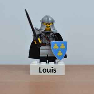 May include: A Lego knight figurine with a silver helmet, armor, and a black cape. The knight holds a black sword and a blue shield with yellow clover symbols. The base reads "Louis".
