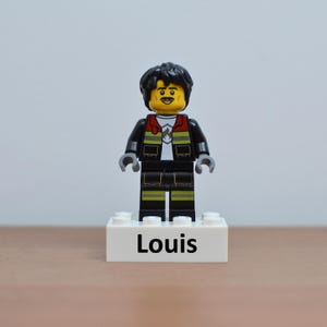 Peut inclure: Une figurine Lego d'un homme aux cheveux noirs et à la moustache, portant une veste noire avec des détails rouges et jaunes sur une chemise blanche. La figurine est sur une base blanche avec le nom "Louis" imprimé.