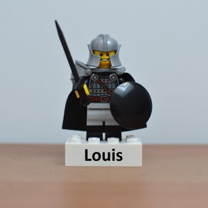 May include: A Lego figurine of a knight in silver armor, holding a black sword and shield. The figurine has a black cape and is standing on a white base with the name "Louis" printed on it.
