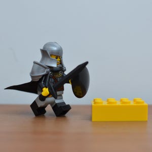 May include: A Lego knight minifigure in silver armor, black cape, and a black sword and shield. The knight is walking towards a yellow Lego brick. The background is a plain, light gray.