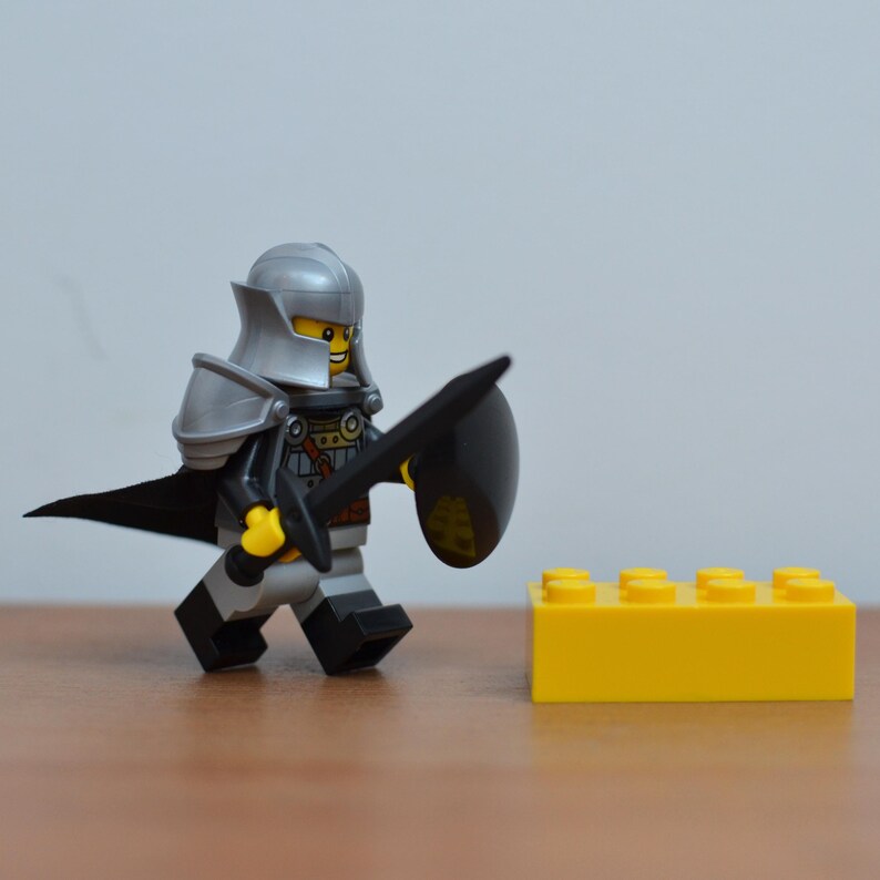 May include: A Lego knight minifigure in silver armor, black cape, and a black sword and shield. The knight is walking towards a yellow Lego brick. The background is a plain, light gray.