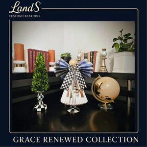 Puede incluir: Una figura decorativa de &aacute;ngel con un lazo y alas a cuadros blancos y negros, un peque&ntilde;o &aacute;rbol verde y un globo dorado sobre una superficie negra. El texto "Grace Renewed Collection" est&aacute; en la parte inferior.
