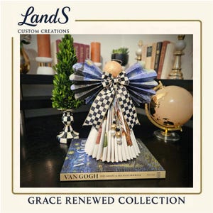 Puede incluir: Una figura decorativa de &aacute;ngel con alas estampadas en azul y negro y un lazo a cuadros. El &aacute;ngel est&aacute; posado sobre un libro titulado "Van Gogh The Artist & His Masterpieces". La figura es parte de la "Grace Renewed Collection". La marca "LandS Custom Creations" es visible.