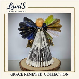 Puede incluir: Una escultura de &aacute;ngel hecha a mano con papel plegado, con una cabeza beige, un halo dorado y alas con detalles azules, verdes y dorados. El cuerpo del &aacute;ngel est&aacute; hecho de papel plegado en blanco y negro. La base es una rodaja de madera. El texto "Grace Renewed Collection".