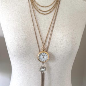 May include: A gold-toned necklace with multiple chains. The focal point is a pocket watch pendant with a white face and black numerals. Below the watch hangs a decorative crystal and tassel.