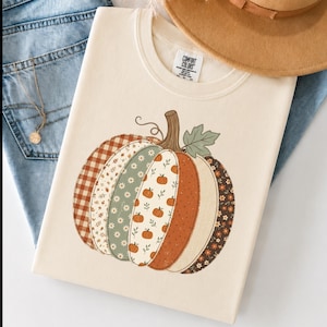 May include: A cream-colored t-shirt featuring a large graphic of a pumpkin composed of vertical segments with various patterns, including brown gingham, white daisies on green, small orange pumpkins, and floral prints, with a brown stem and green leaf.