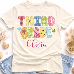 May include: A cream-colored t-shirt featuring the text "THIRD GRADE" in colorful, patterned, block-style letters decorated with small hearts, stars, and a smiley face. Below the text, the name "Olivia" is printed in a purple cursive font.