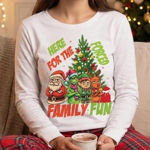 May include: White long-sleeve shirt with a Christmas-themed graphic. The graphic features Santa Claus, the Grinch, an elf, and a reindeer in front of a Christmas tree. The text reads "Here for the Family Fun Forced."