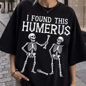 May include: A black t-shirt featuring a white graphic of two skeletons and the text I FOUND THIS HUMERUS. One skeleton holds a bone while the other stands nearby, with a detached arm bone lying on the ground below the skeletons in the center.