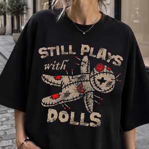 May include: A black t-shirt featuring a graphic of a beige voodoo doll with red accents and stitches. The text "STILL PLAYS with DOLLS" is printed in a distressed beige font above and below the doll illustration on the front of the garment.