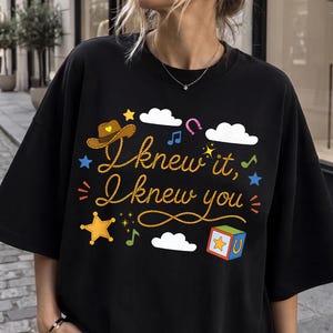 May include: A black t-shirt featuring a graphic print with the text "I knew it, I knew you" in a rope-style font. The design includes a brown cowboy hat, white clouds, a yellow star, a colorful block, musical notes, and a pink horseshoe.