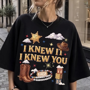 May include: A black short-sleeved t-shirt featuring a graphic print with the text "I KNEW IT I KNEW YOU". The design includes a gold sheriff star labelled "INFINITY AND BEYOND", a brown cowboy hat, brown cowboy boots, a beaded bracelet, and blocks.