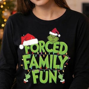 May include: Black long-sleeve shirt with the text "FORCED FAMILY FUN" in green, featuring a Grinch-like character and Santa hats. The person is wearing red and green plaid pajama pants. Christmas-themed apparel.