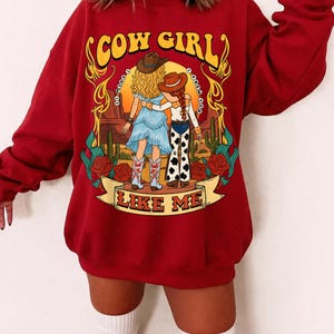 May include: A dark red long-sleeve sweatshirt featuring a graphic print of two figures in a desert landscape. The text reads COW GIRL and LIKE ME. The illustration includes cacti, red roses, and yellow decorative flourishes surrounding the figures.