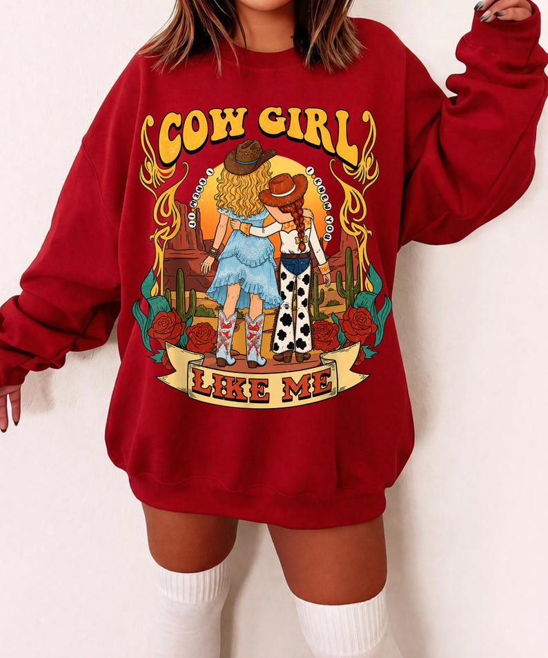 May include: A dark red long-sleeve sweatshirt featuring a graphic print of two figures in a desert landscape. The text reads COW GIRL and LIKE ME. The illustration includes cacti, red roses, and yellow decorative flourishes surrounding the figures.