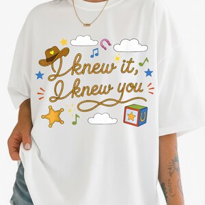 May include: A white t-shirt featuring a graphic print with the text "I knew it, I knew you" written in a rope-style font. The design includes a brown cowboy hat, a yellow sheriff star, a colorful toy block, clouds, stars, and musical notes.
