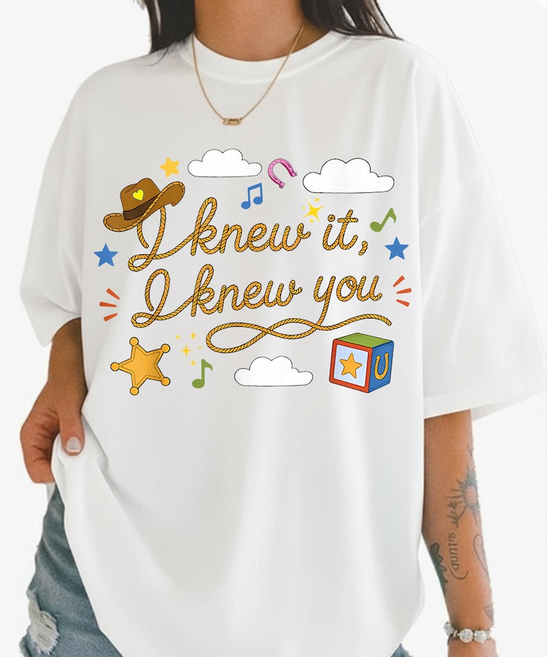 May include: A white t-shirt featuring a graphic print with the text "I knew it, I knew you" written in a rope-style font. The design includes a brown cowboy hat, a yellow sheriff star, a colorful toy block, clouds, stars, and musical notes.