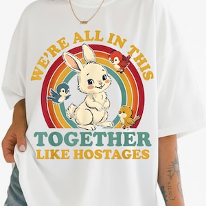 May include: White t-shirt with a cartoon rabbit and birds set against a rainbow. The text reads "WE'RE ALL IN THIS TOGETHER LIKE HOSTAGES" in yellow and teal. The design is centered on the shirt.