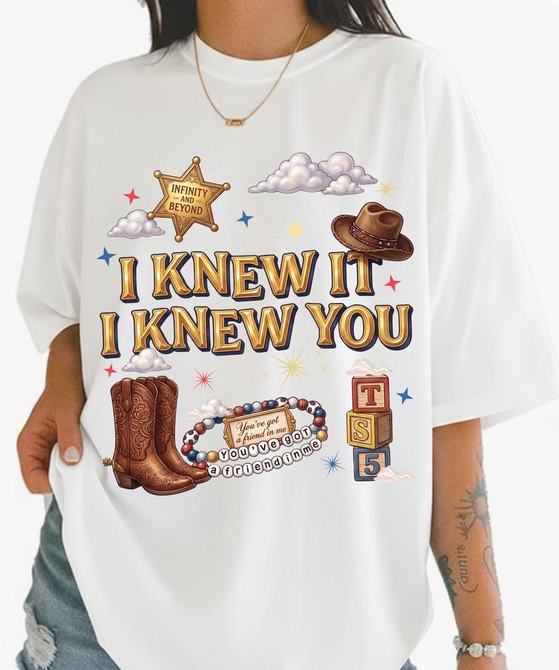 May include: A white t-shirt featuring the text I KNEW IT I KNEW YOU in gold lettering. The graphic includes a sheriff badge with the text INFINITY AND BEYOND, a brown cowboy hat, brown cowboy boots, a beaded bracelet, and three stacked blocks.