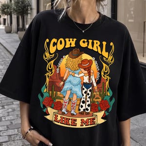 May include: A black t-shirt featuring a graphic illustration of two figures in cowboy hats standing in a desert landscape with cacti and red roses. The text reads COW GIRL at the top and I KNEW YOU LIKE ME at the bottom in yellow and red lettering.
