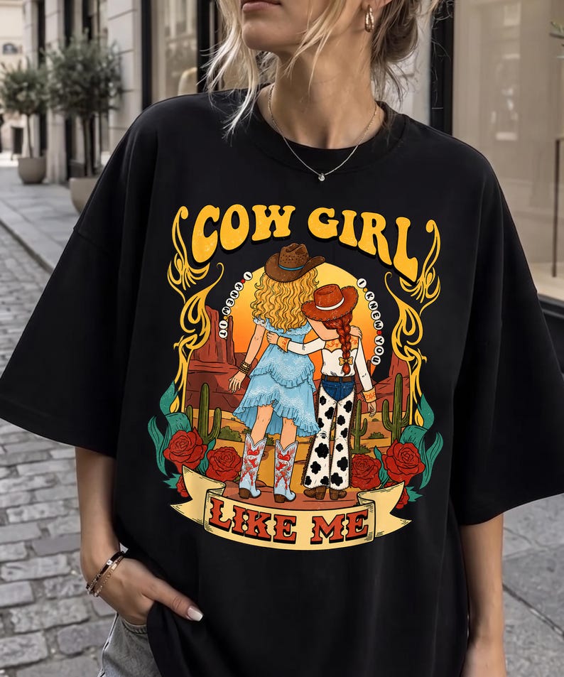 May include: A black t-shirt featuring a graphic illustration of two figures in cowboy hats standing in a desert landscape with cacti and red roses. The text reads COW GIRL at the top and I KNEW YOU LIKE ME at the bottom in yellow and red lettering.