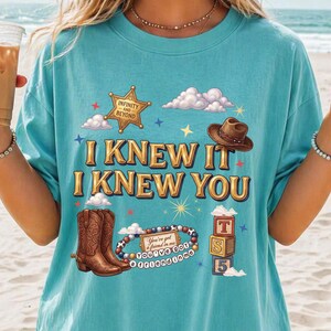 May include: A turquoise t-shirt featuring a graphic print with the text "I KNEW IT I KNEW YOU". The design includes a gold sheriff badge labeled "INFINITY AND BEYOND", a brown cowboy hat, brown cowboy boots, a beaded bracelet, and alphabet blocks.