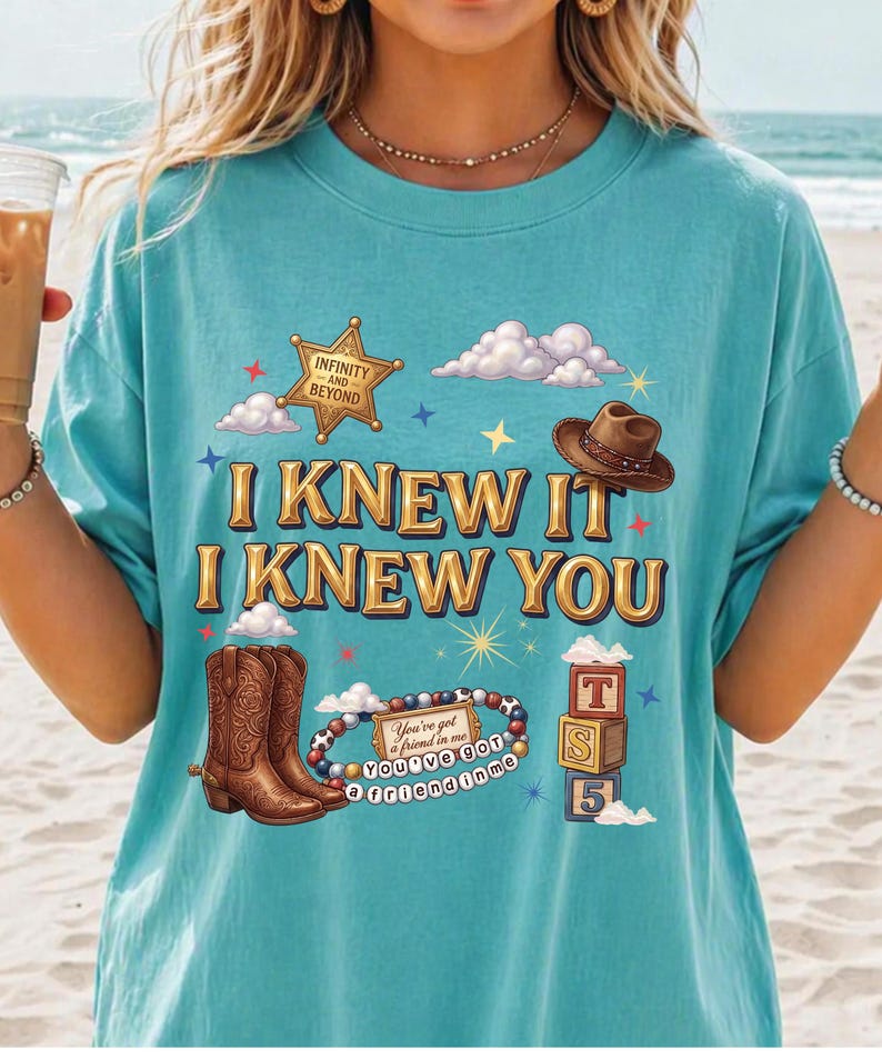 May include: A turquoise t-shirt featuring a graphic print with the text "I KNEW IT I KNEW YOU". The design includes a gold sheriff badge labeled "INFINITY AND BEYOND", a brown cowboy hat, brown cowboy boots, a beaded bracelet, and alphabet blocks.