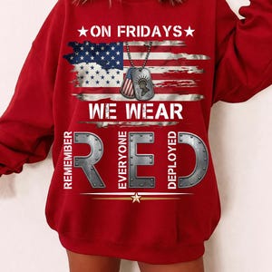 May include: A red sweatshirt featuring the text "ON FRIDAYS WE WEAR RED" and an American flag design. The design includes military dog tags. The words "REMEMBER EVERYONE DEPLOYED" are also printed on the sweatshirt.