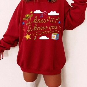 May include: A deep red long-sleeve sweatshirt featuring the yellow text "I knew it, I knew you" in a cursive font. The design includes a brown cowboy hat, white clouds, a colorful toy block, a yellow star, musical notes, and blue stars.