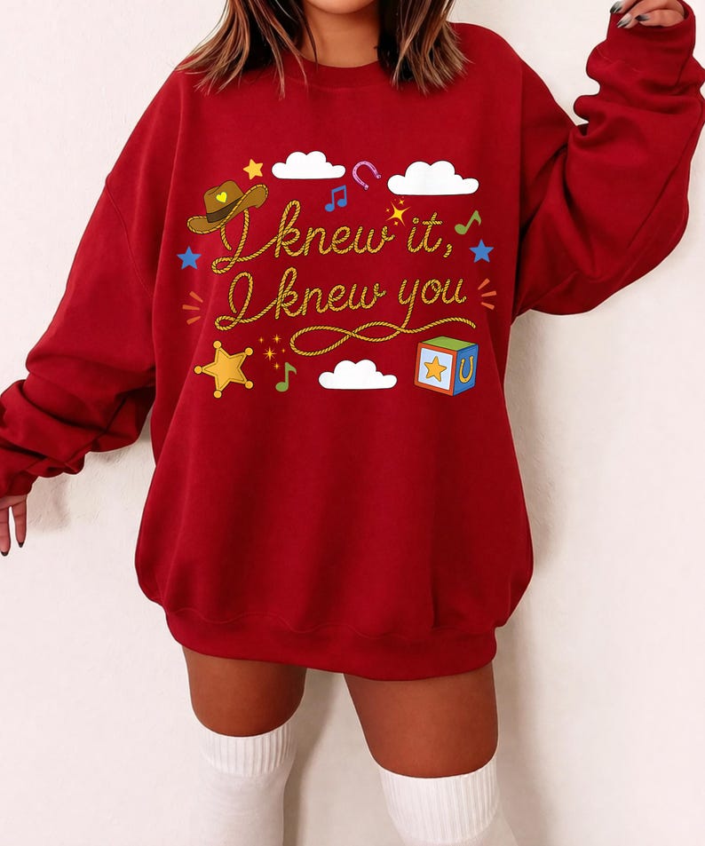 May include: A deep red long-sleeve sweatshirt featuring the yellow text "I knew it, I knew you" in a cursive font. The design includes a brown cowboy hat, white clouds, a colorful toy block, a yellow star, musical notes, and blue stars.