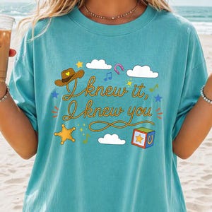 May include: A teal t-shirt featuring a graphic print with the text "I knew it, I knew you" written in a rope-style font. The design includes a brown cowboy hat, white clouds, a yellow star, a colorful block, a horseshoe, and musical notes.