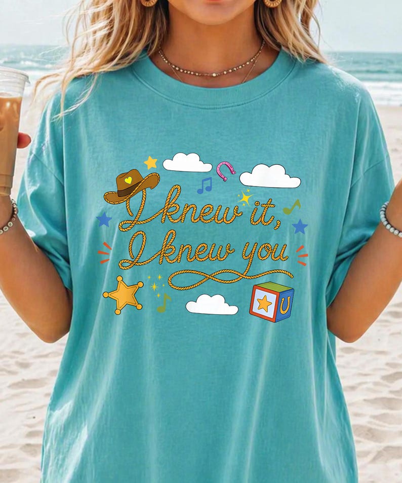 May include: A teal t-shirt featuring a graphic print with the text "I knew it, I knew you" written in a rope-style font. The design includes a brown cowboy hat, white clouds, a yellow star, a colorful block, a horseshoe, and musical notes.