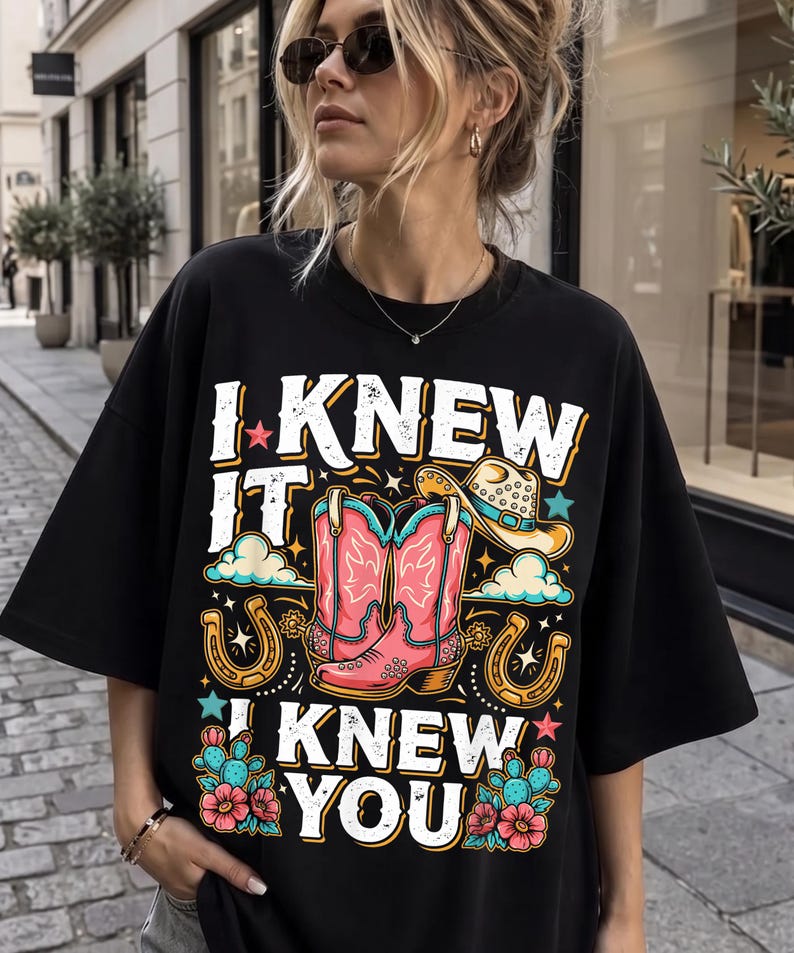 May include: A black t-shirt featuring a graphic print with the text I KNEW IT I KNEW YOU. The design includes pink cowboy boots, a tan cowboy hat, two gold horseshoes, blue clouds, small stars, and green cacti with pink flowers.