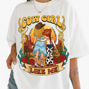 May include: A white t-shirt featuring a graphic illustration of two figures in cowboy hats standing in a desert landscape with cacti and red roses. The text reads COW GIRL at the top and LIKE ME at the bottom, with I KNEW IT and I KNEW YOU in circles.