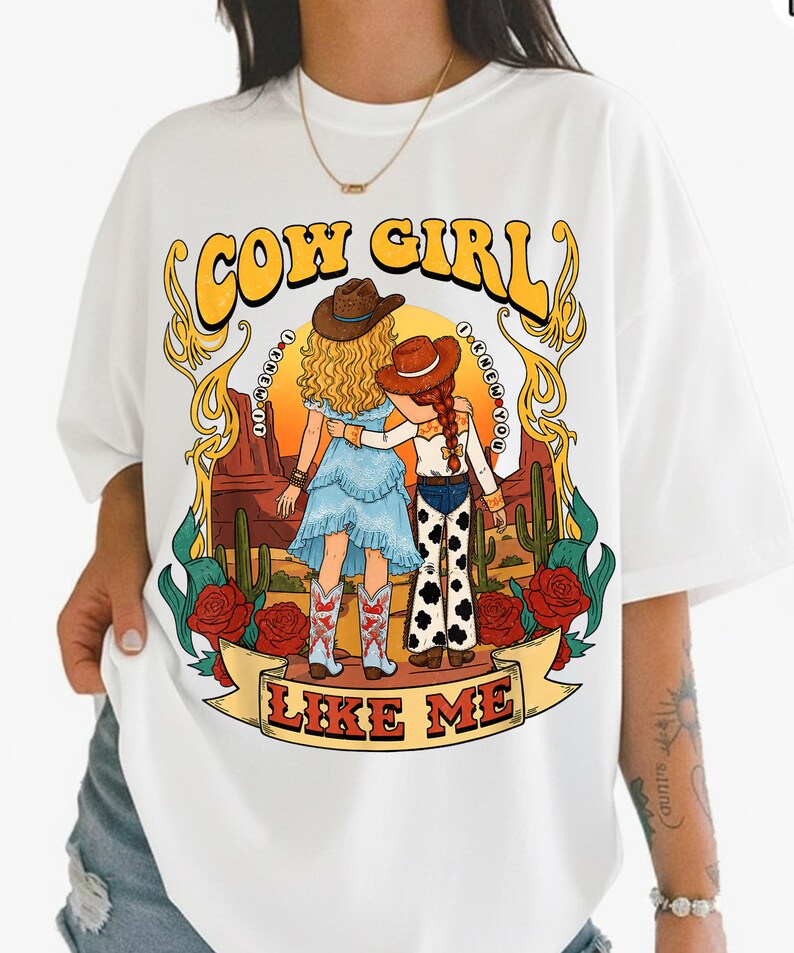 May include: A white t-shirt featuring a graphic illustration of two figures in cowboy hats standing in a desert landscape with cacti and red roses. The text reads COW GIRL at the top and LIKE ME at the bottom, with I KNEW IT and I KNEW YOU in circles.