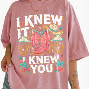 May include: A dusty pink short-sleeve t-shirt featuring a graphic print with the text I KNEW IT I KNEW YOU. The design includes a pair of pink cowboy boots, a cowboy hat, two horseshoes, small stars, clouds, and teal cacti with pink flowers.