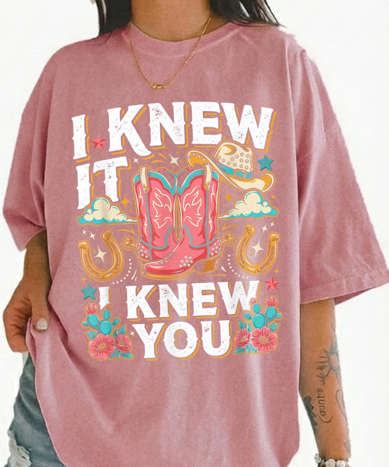 May include: A dusty pink short-sleeve t-shirt featuring a graphic print with the text I KNEW IT I KNEW YOU. The design includes a pair of pink cowboy boots, a cowboy hat, two horseshoes, small stars, clouds, and teal cacti with pink flowers.