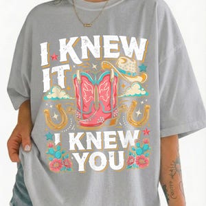 May include: A light gray t-shirt featuring a graphic print with the text "I KNEW IT I KNEW YOU" in white. The design includes a pair of pink cowboy boots, a tan cowboy hat, two gold horseshoes, teal cacti, and pink flowers with small stars.