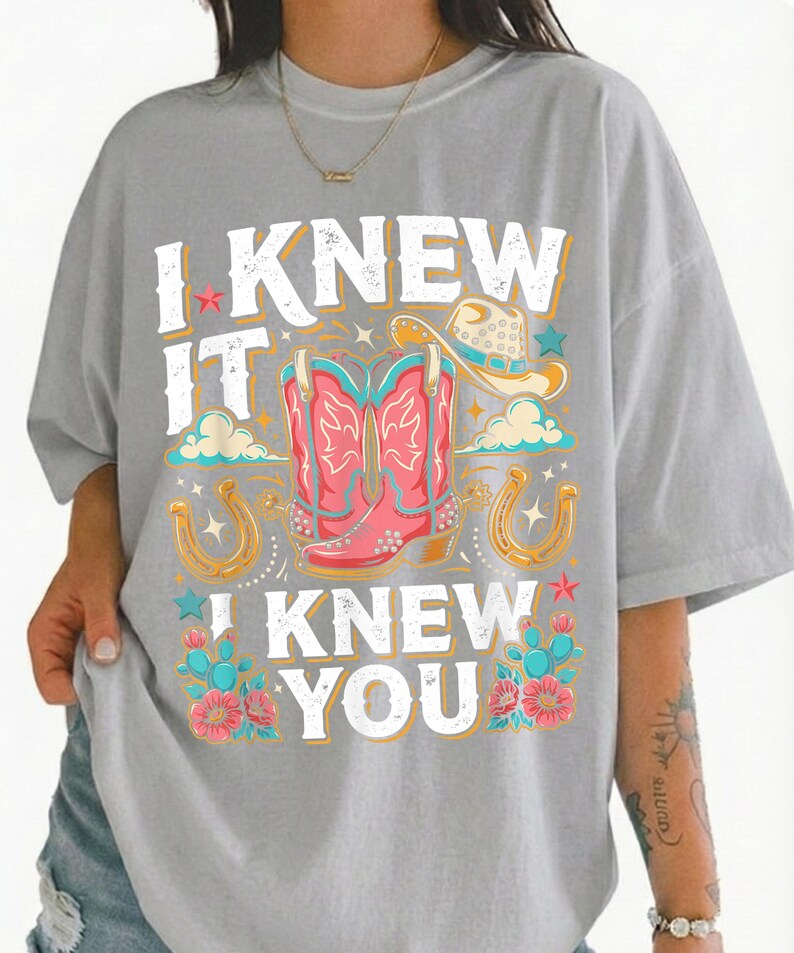 May include: A light gray t-shirt featuring a graphic print with the text "I KNEW IT I KNEW YOU" in white. The design includes a pair of pink cowboy boots, a tan cowboy hat, two gold horseshoes, teal cacti, and pink flowers with small stars.